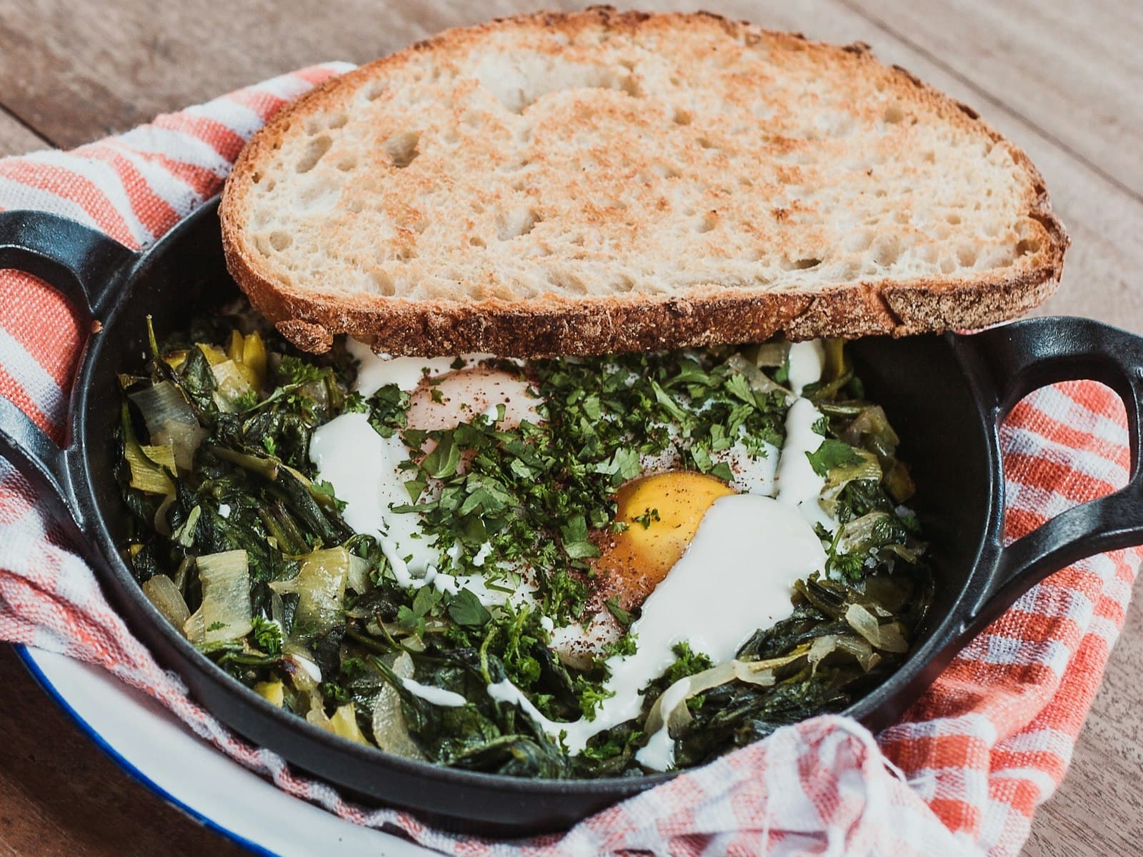 Gusseiserne Pfanne mit Spinat, Lauch und gestocktem Ei, beträufelt mit Joghurt und bestreut mit gehackten Kräutern, darauf eine Scheibe geröstetes Brot