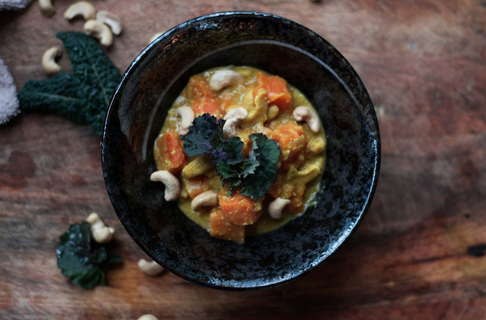 Dunkle Schale mit cremigem Kürbis-Curry, Kürbiswürfeln und Cashewkernen, garniert mit einem Grünkohlblatt, auf einem Holzbrett